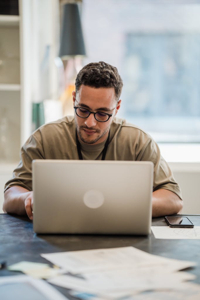 Man in glasses concentrating on work at desk with laptop in bright office setting.